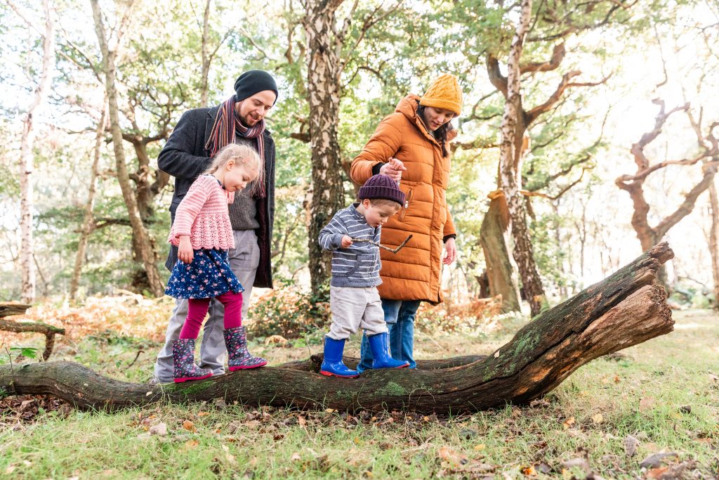 „Zwei Erwachsene unterstützen zwei Kinder beim Balancieren über einen Baumstamm im Wald.“